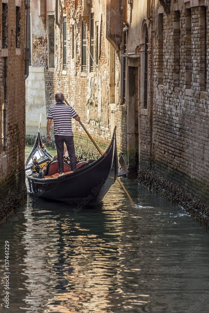 Venetian gondolier navigates with his gondola in a narrow canal in the historic center of Venice, Italy