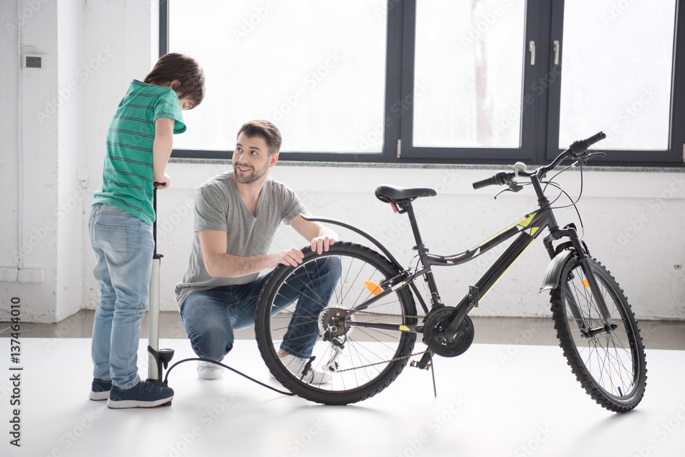 Happy father looking at son inflating bicycle tire with pump indoors Happy father looking at son inflating bicycle tire with pump indoors