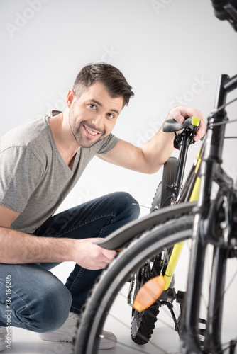 Young handsome man crouching while checking bicycle and smiling at camera