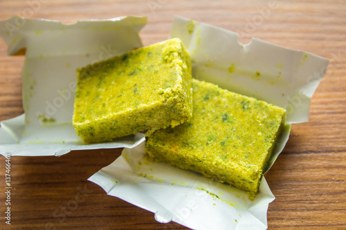Dehydrated bouillon stock cube salty meat and vegetables aromatic yellow spice, ingredient single whole condiment portion wrapped, open in paper pack on wooden background