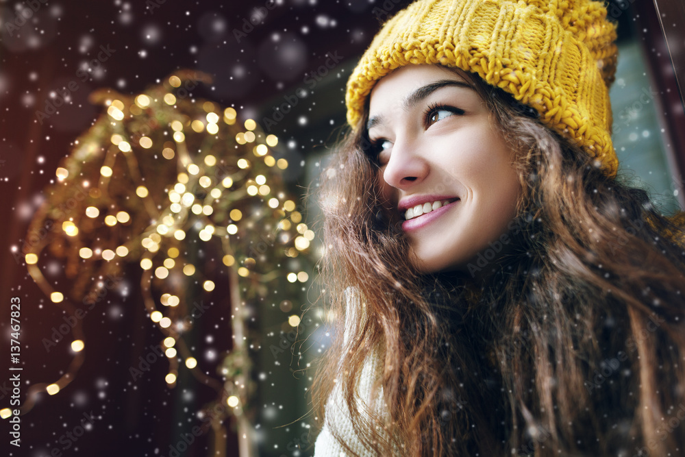 Outdoor close up portrait of young beautiful happy smiling girl posing on street. Model looking aside, wearing stylish winter hat. Snowfall, festive garland. City lifestyle. Copy, empty space for text Outdoor close up portrait of young beautiful happy smiling girl posing on street. Model looking aside, wearing stylish winter hat. Snowfall, festive garland. City lifestyle. Copy, empty space for text