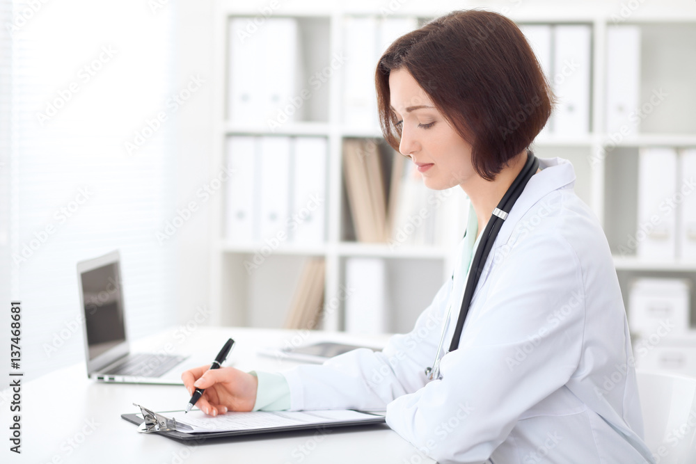 Young brunette female doctor sitting at the table and working at hospital office. Health care, insurance and help concept. Physician ready to examine patient Young brunette female doctor sitting at the table and working at hospital office. Health care, insurance and help concept. Physician ready to examine patient