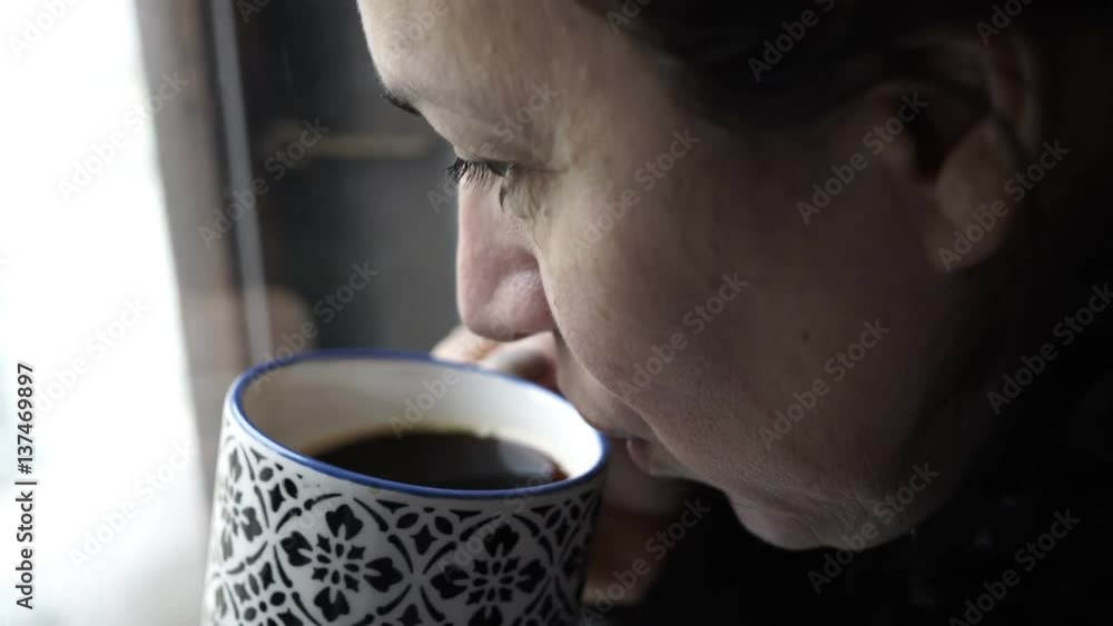 Handheld shot of a woman looking out of a window on a cold morning and drinking a cup of coffee. Handheld shot of a woman looking out of a window on a cold morning and drinking a cup of coffee.