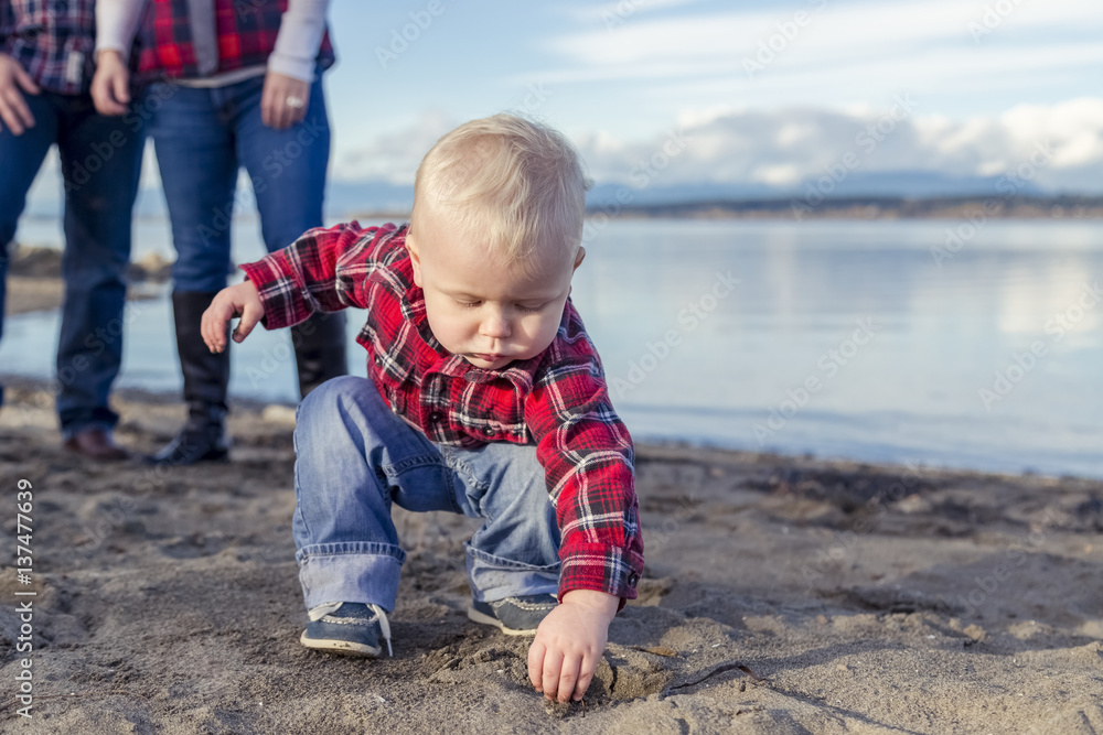 A young toddler gets his hands dirty playing with sand on the beach while his parents look on from the background; Surrey, British Columbia, Canada A young toddler gets his hands dirty playing with sand on the beach while his parents look on from the background; Surrey, British Columbia, Canada