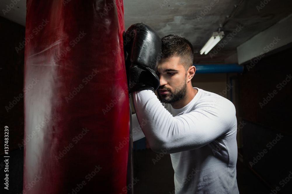 Portrait of tired young man leaning on punching bag after hard workout at the gym