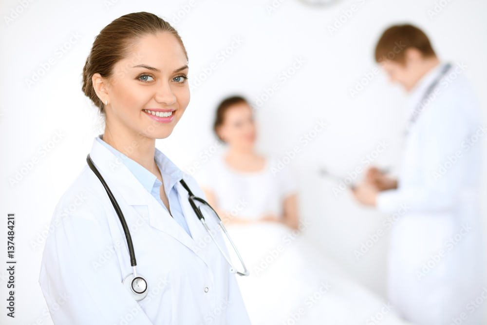 Cheerful smiling female doctor on the background with physician and his patient in the bed. High level and quality of medical service concept. Cheerful smiling female doctor on the background with physician and his patient in the bed. High level and quality of medical service concept.