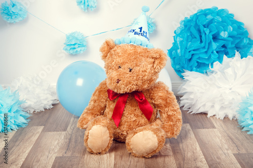 Festive background decoration for first year birthday celebration with little bear toy sitting on floor in studio, balloons, paper flowers and presents gift boxes, toned with filters