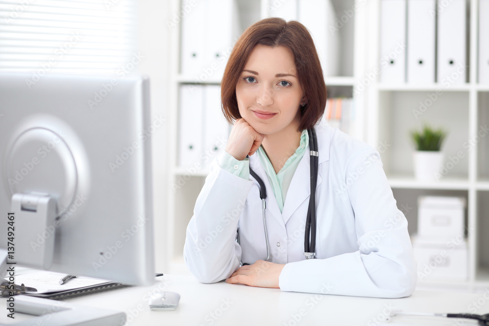 Young brunette female doctor sitting at a desk and working on the computer at the hospital office. Health care, insurance and help concept. Physician ready to examine patient Young brunette female doctor sitting at a desk and working on the computer at the hospital office. Health care, insurance and help concept. Physician ready to examine patient