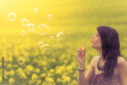 Beautiful woman blowing funny soap bubbles in summer nature. The genuine girl has fun playing in a yellow green meadow and is enjoying her youth