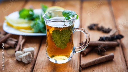 Clear glass of hot black herbal tea with lemon, mint and Melissa leaves on light rustic wooden table. Summer, Autumn, winter drink. Cinnamon and cane sugar for decoration.
