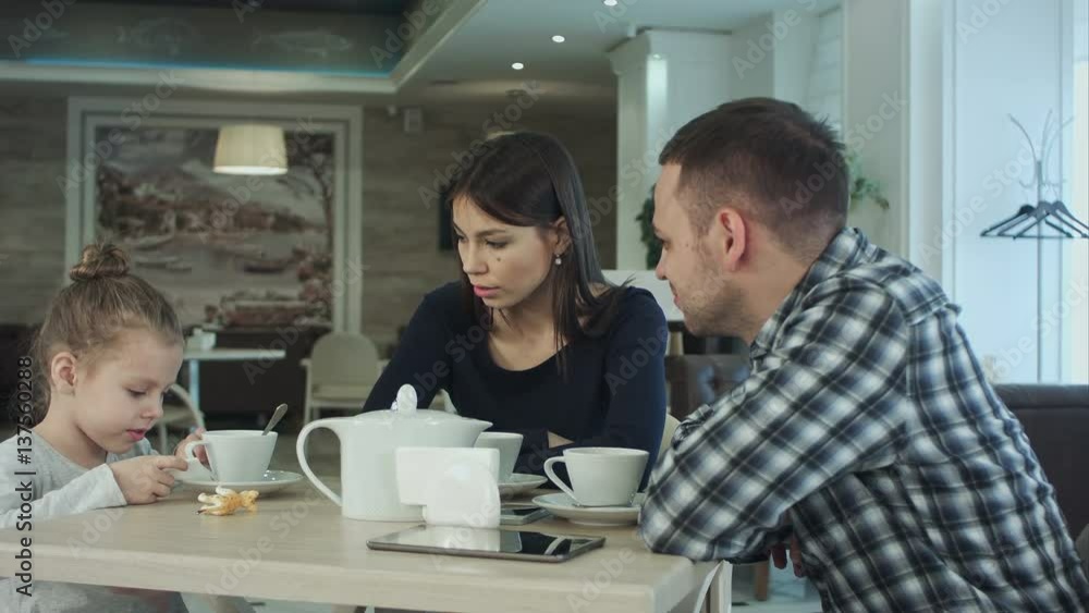 Family Enjoying tea In Cafe Together. Father and mother talking looking at their daughter. Family Enjoying tea In Cafe Together. Father and mother talking looking at their daughter.