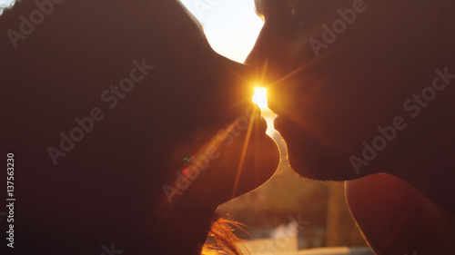 Romantic young beautiful couple in love looking each other and kissing on a sunset with sun shining bright behind them on a horizon.