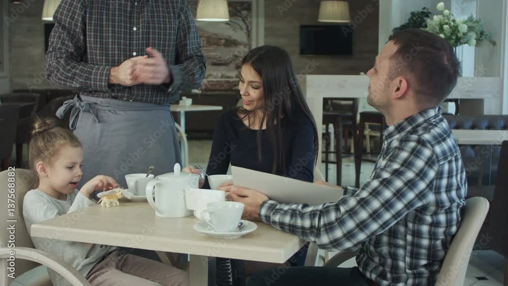 Waiter giving menu to happy family at cafe. Father consulting with him. Waiter giving menu to happy family at cafe. Father consulting with him.