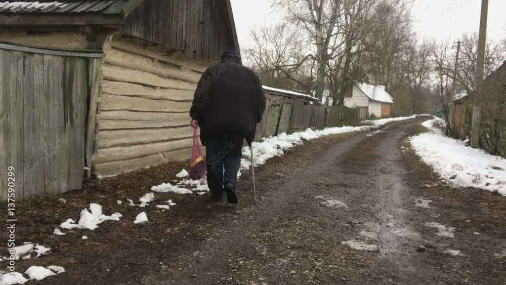 Senior Ukrainian peasant with walking stick carrying string-bag moving on a winter street in Ukrainian village Senior Ukrainian peasant with walking stick carrying string-bag moving on a winter street in Ukrainian village
