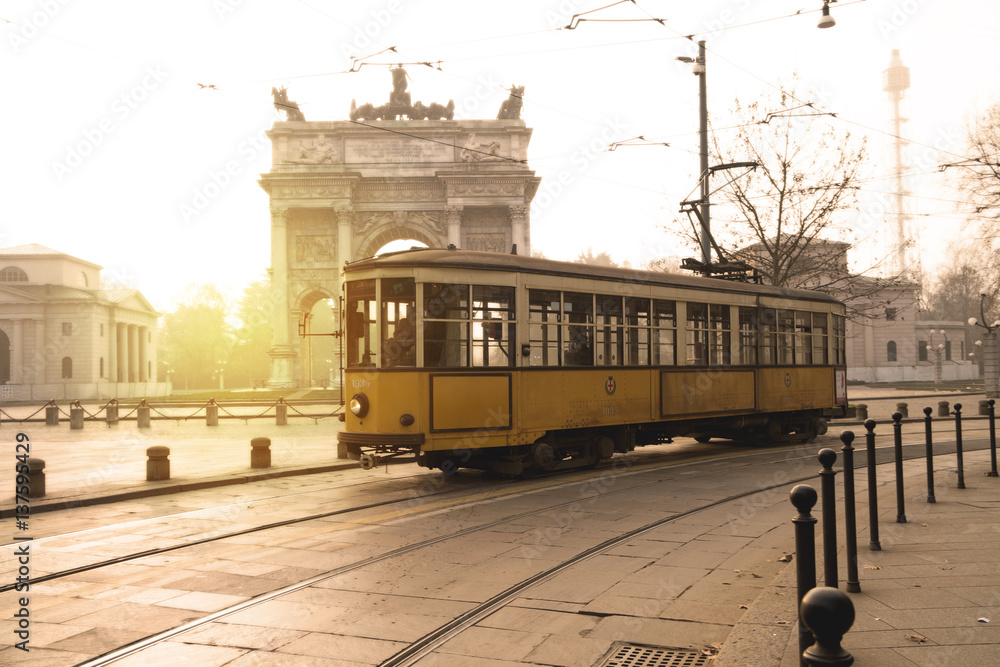 Fototapeta premium Stary żółty tramwaj przejeżdża o świcie przed Łukiem Pokoju w parku Sempione w Mediolanie we Włoszech.