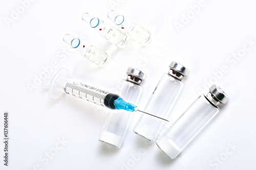 Medical syringe, three ampules with saline and vials with dry human hormone drug powder. Syringe with bare needle lying on vials. Macro image. White background, shadows.