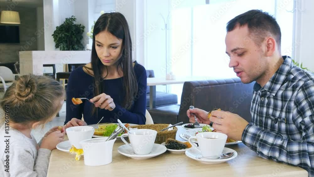 Young lovely parents feeding their blond daughter vegetable salad in cafe. Young lovely parents feeding their blond daughter vegetable salad in cafe.