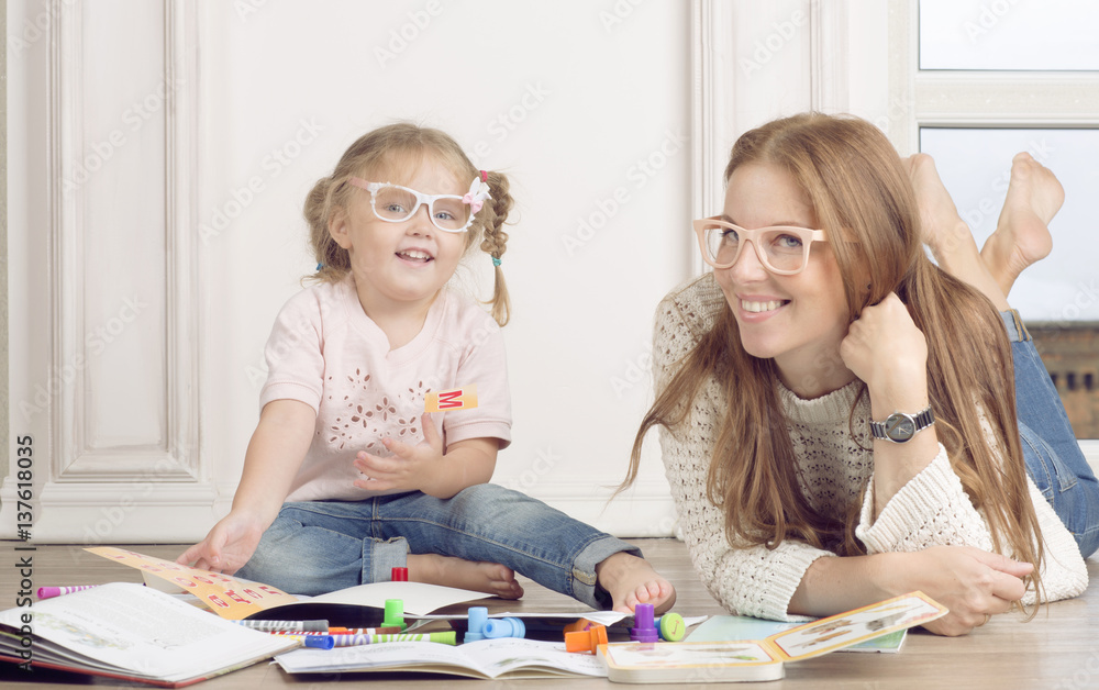 The daughter and mother sit on the floor and draw. The woman helps to the child to draw with markers. The daughter and mother sit on the floor and draw. The woman helps to the child to draw with markers.