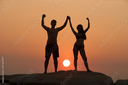 Positive couple holding hand up and expressing gladness while standing on stone in Sunset.