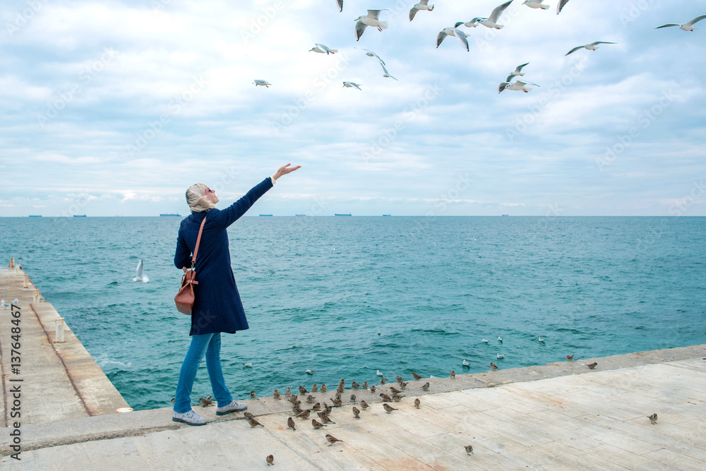 blonde woman feeding seagulls in cloudy autumn day on the sea coast 