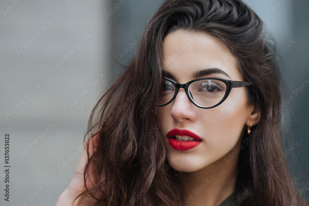 Fototapeta premium Fashion consept: beatiful young girl with long hair, glasses, red lips standing near modern wall wearing in green suit and grey jeans. She wanted talking on phone.