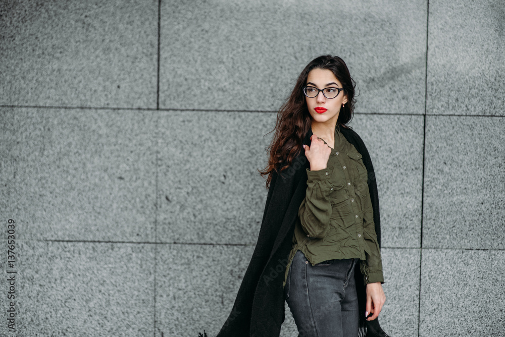 Fashion consept: beatiful young girl with long hair, glasses, red lips standing near modern wall wearing in green suit and grey jeans. She wanted talking on phone. Fashion consept: beatiful young girl with long hair, glasses, red lips standing near modern wall wearing in green suit and grey jeans. She wanted talking on phone.