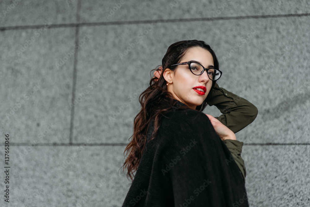 Naklejka premium Fashion consept: beatiful young girl with long hair, glasses, red lips standing near modern wall wearing in green suit and grey jeans. She wanted talking on phone.