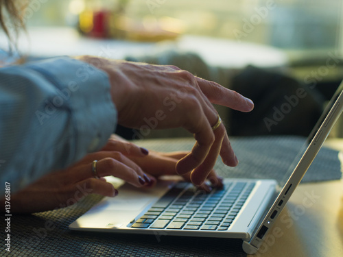 Woman's hands typing on laptop keyboard in interior, side view of businessmans using computer in cafe.