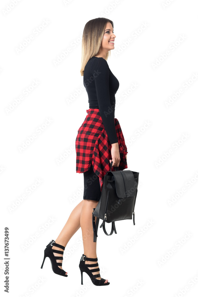Happy young street style woman in stiletto and black dress walking and smiling side view. Full body length isolated over white studio background. Happy young street style woman in stiletto and black dress walking and smiling side view. Full body length isolated over white studio background.