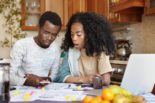 African male in glasses calculating family expenses holding cell phone, his surprised wife with curly hair looking at screen in shock, keeping mouth wide open, astonished with amount of unpaid bills