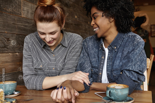 Love and relationships. Cheerful African lesbian with braces and Afro hairstyle holding hands with her cute redhead girlfriend, talking to each other at cafe, sharing happy and sweet moments together photo