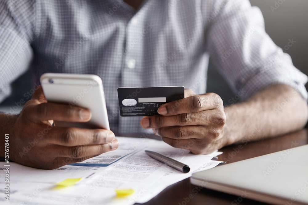 Cropped shot of African-American male paying bill at restaurant with online payment technology via internet, using free wi-fi during breakfast, sitting at table with plastic card and smart phone Cropped shot of African-American male paying bill at restaurant with online payment technology via internet, using free wi-fi during breakfast, sitting at table with plastic card and smart phone