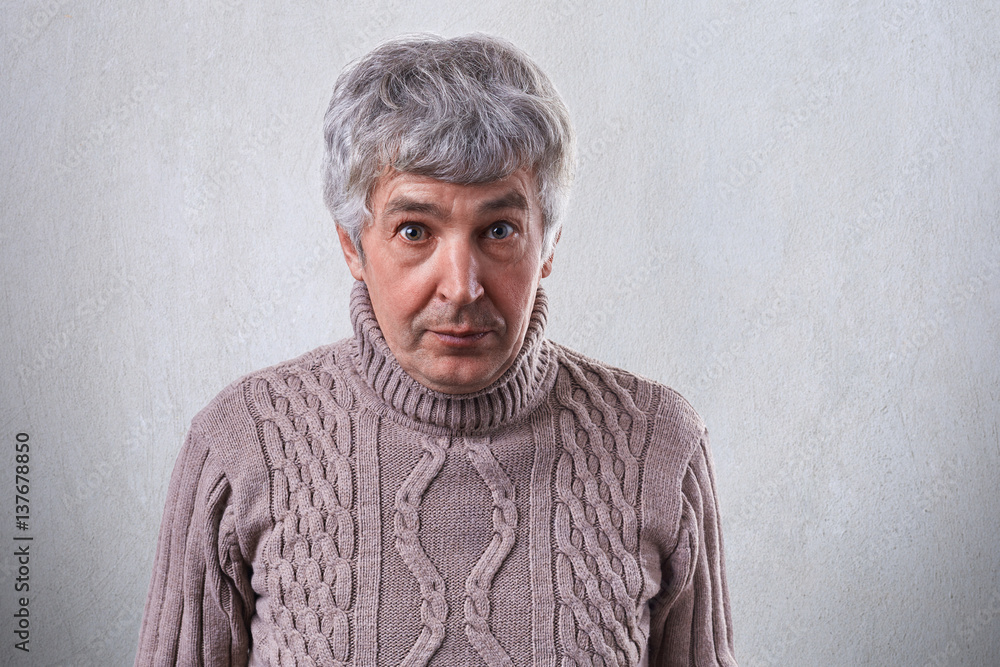 A horizontal portrait of senior handsome man standing against white wall wearing sweater looking with astonishment into camera. People, emotions, feelings, astonishment, wonder, amazement A horizontal portrait of senior handsome man standing against white wall wearing sweater looking with astonishment into camera. People, emotions, feelings, astonishment, wonder, amazement