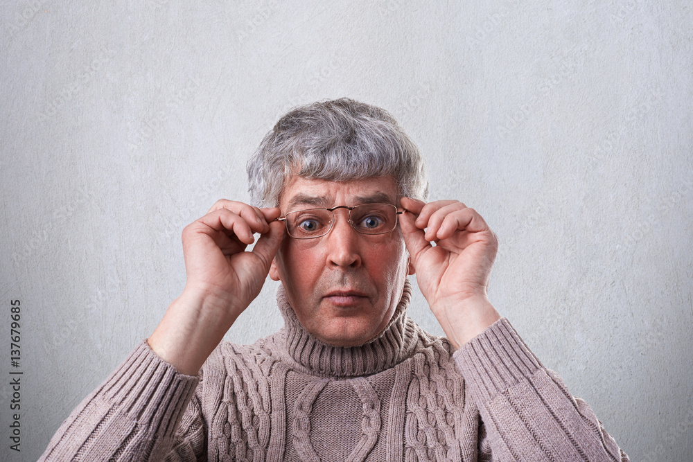A close-up of astonished senior man wearing glasses and sweater holding his hands on the frames of glasses looking with wide opened eyes into camera. A surprised elderly man over white background A close-up of astonished senior man wearing glasses and sweater holding his hands on the frames of glasses looking with wide opened eyes into camera. A surprised elderly man over white background