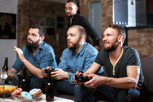 Men with a beard sitting on the couch at home with beer and chips with joysticks in hand playing computer video games. The concept of friendship, technology and weekend