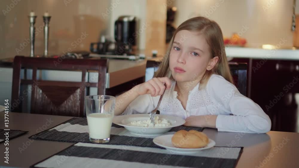 Girl 8 years old do not want eat cottage cheese. Sad girl sitting in the kitchen Girl 8 years old do not want eat cottage cheese. Sad girl sitting in the kitchen