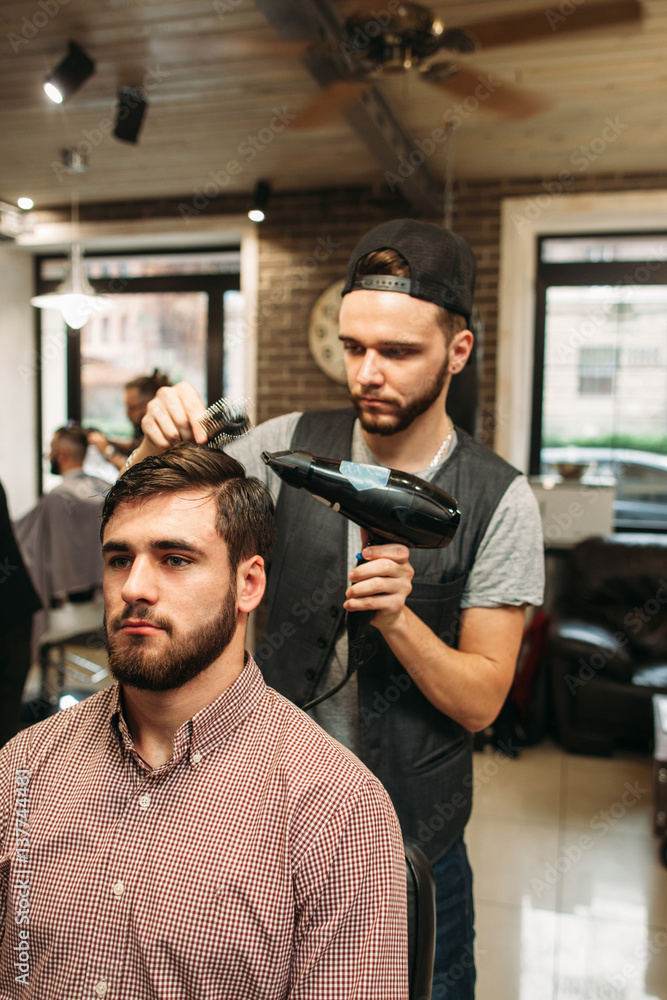 Stylist making modern hairdo for young man. Successful businessman sitting at barber shop and dry hair. Style, beauty, modern life concept Stylist making modern hairdo for young man. Successful businessman sitting at barber shop and dry hair. Style, beauty, modern life concept