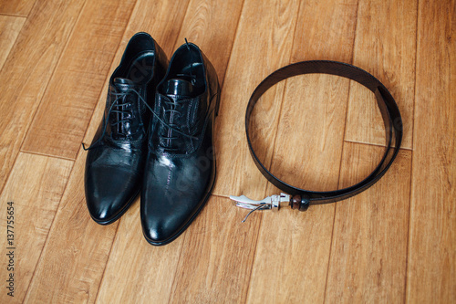 Men's classic black leather shoes and a leather belt with a buckle on wooden background.