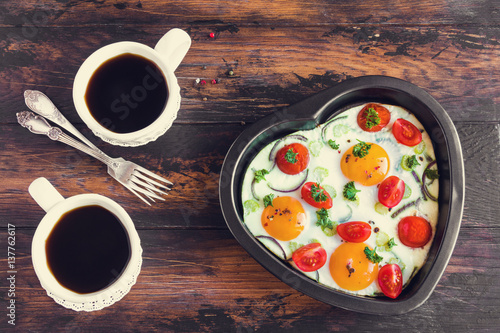 Romantic breakfast for Valentine day or anniversary, fried eggs with tomatoes, celery, onion and parsley in heart shaped baking dish and black coffee. Rustic wooden table, top view.