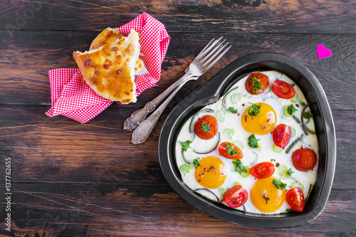 Romantic breakfast for Valentine day or anniversary, fried eggs with tomatoes, celery, onion and parsley in black heart shaped baking dish. Rustic wooden table, bread and vintage forks, top view.