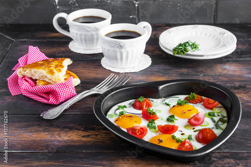 Romantic breakfast for Valentine day or anniversary, fried eggs with tomatoes, celery, onion and parsley in heart shaped baking dish and black coffee. Rustic wooden table, fresh vegetables and bread.