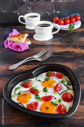 Romantic breakfast for Valentine day or anniversary, fried eggs with tomatoes, celery, onion and parsley in heart shaped baking dish and black coffee. Rustic wooden table, fresh vegetables and bread.