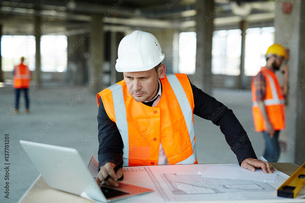 © pressmaster - Portrait of development supervisor wearing protective vest and helmet over formal suit  proofing blueprints using laptop computer inside unfinished building
