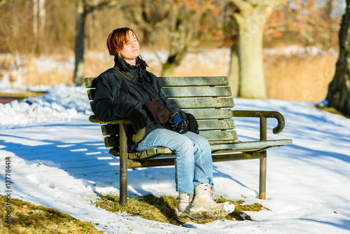 Fotografie Young adult female dressed in black and blue, basking in sunshine on park bench in winter or early spring
