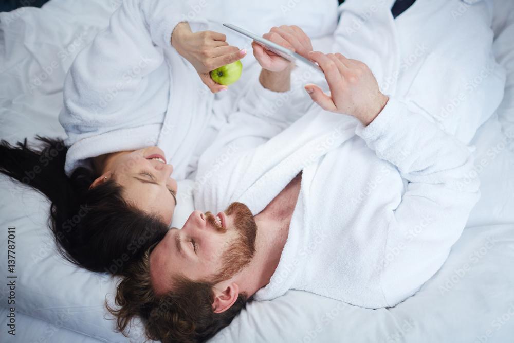 Modern couple in bathrobes using touchpad in bed Modern couple in bathrobes using touchpad in bed