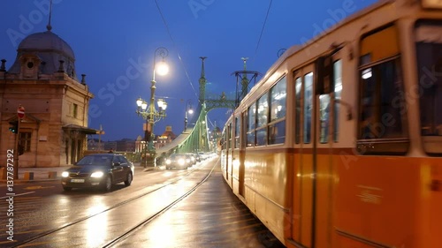 Nice view of  the Liberty Bridge in Budapest night. Beautiful lighting. City lights reflected on a wet road. On the bridge are people walking, riding cars, tram