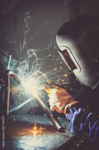 Industrial worker welding round pipe on a work table, producing smoke, sparks and colorful reflections
