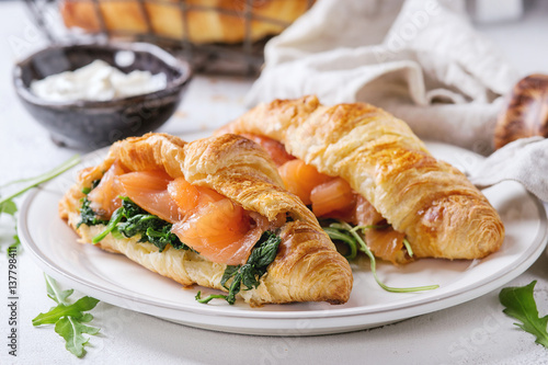Croissant with smoked salted salmon, spinach and arugula served on white plate with bowl of cream cheese and coffee maker over white and gray concrete background. Close up