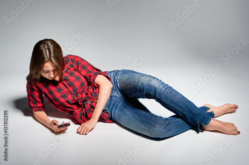 Lovely girl wearing jeans and red checked shirt lying barefooted at floor texting on her cell phone, studio shot
