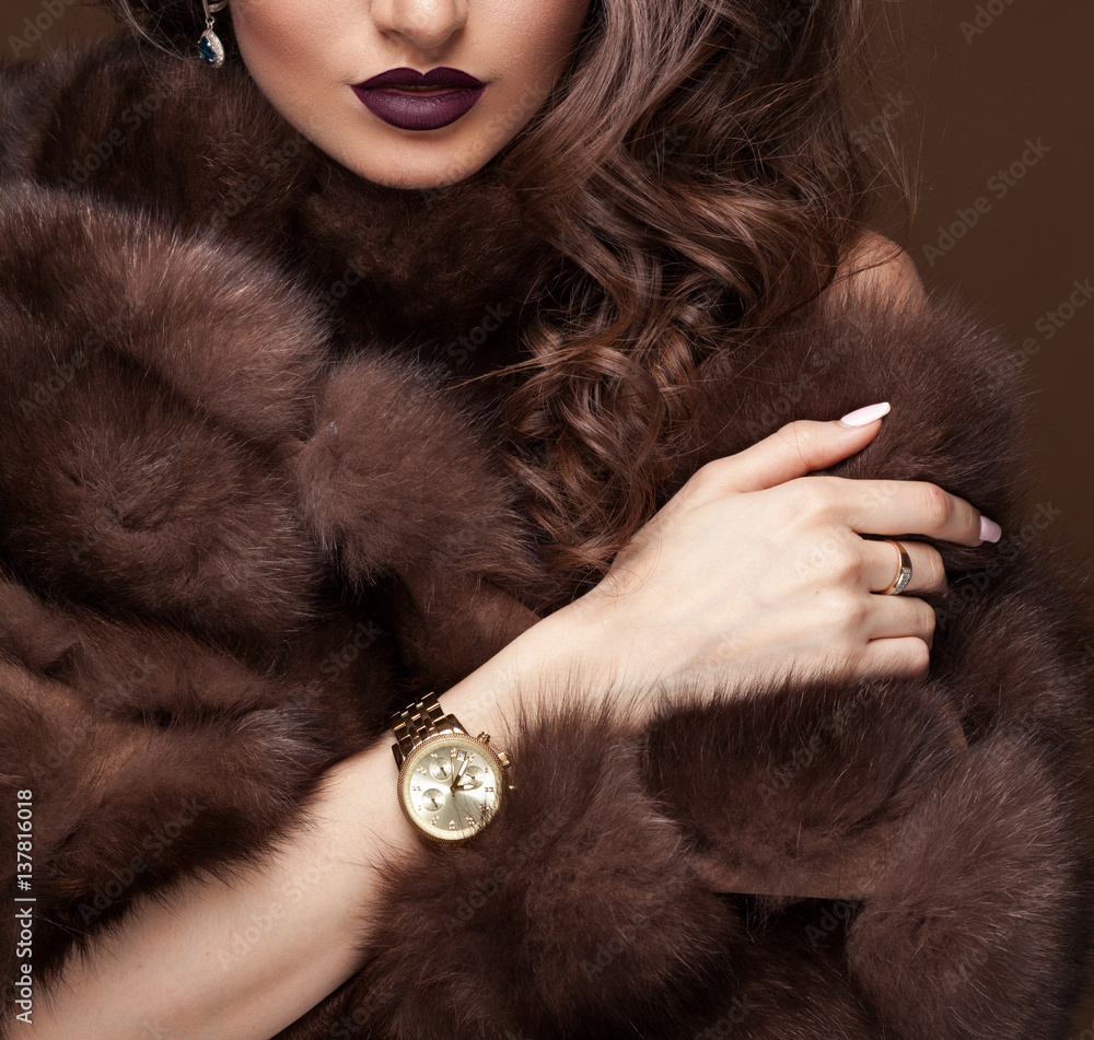 Naklejka premium Luxury elegant woman in fur coat, golden earrings and watches. Glamour beautiful look. Rich lady over dark chocolat background.